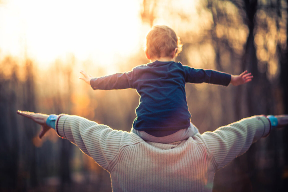 Picture of a grandfather playing with his three year old grandson in the park. The old man is wearing a khaki sweater and a blue shirt with long sleeves. The little boy is wearing a jacket, hat and grey sweatpants. They are smiling and having fun. In the background are trees and yellow leaves. The Lens flare is between them.